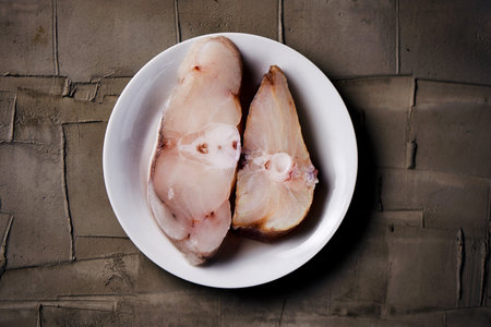 high angle view of some slices of raw monkfish on a white ceramic plate on a gray textured concrete...