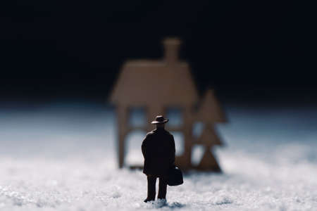 closeup of a miniature man carrying a suitcase on the snow in front of the silhouette of a house, in the backgroundの写真素材