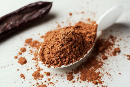 closeup of a pile of carob powder on a white ceramic spoon and a ripe carob pod on a white tableの写真素材