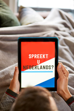 closeup of a young caucasian man, lying comfortably in a sofa wrapped in a warming gray blanket, having his tablet in his hands with the text do you speak dutch in its screenの写真素材