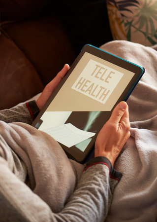 a young caucasian man, lying comfortably in a sofa wrapped in a warming gray blanket, has his tablet in his hands with the text telehealth on its screenの写真素材