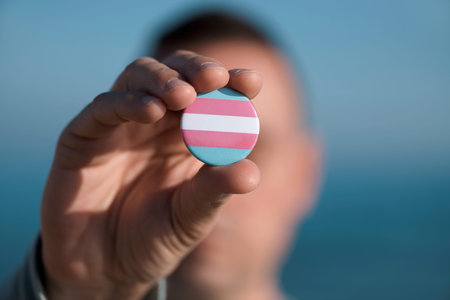 closeup of a young caucasian person showing a pin button patterned with a transgender pride flag in...