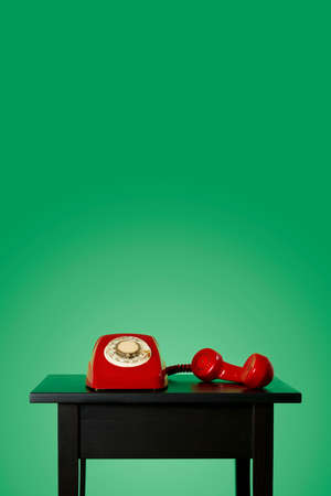 red landline rotary dial telephone, with its handset off the hook on a black wooden table, on a green background with some blank space on topの写真素材