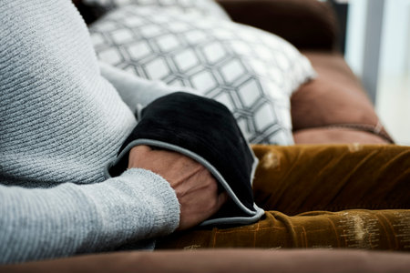 a young caucasian man uses a black electric hot water bag to warm his hands, while is sitting in a...