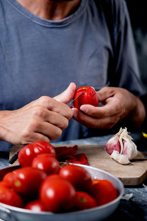 closeup of a young caucasian man, wearing a gray t-shirt, peeling a scalded tomato with a knife at a kitchen table or countertop next to a head of garlicの写真素材