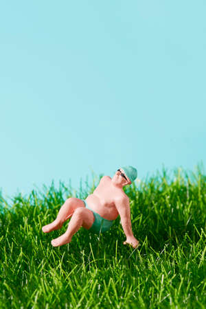 closeup of a miniature man in swimsuit relaxing sitting on the grass, against a blue background with some blank space on topの写真素材