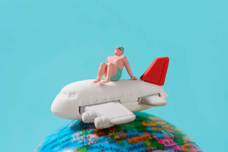 closeup of a miniature man, in swimsuit, sitting on top of an airplane and a terrestrial globe, against a blue background simulating the skyの写真素材