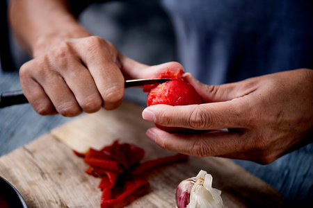 a young caucasian man, in a blue t-shirt, peels a scalded tomato with a knife at a kitchen table or...