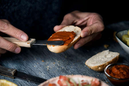 closeup of a young caucasian man preparing a vegan appetizer, by spreading a vegan version of the spanish sobrasada, made with sun-dried tomato and almonds, on a toasted halved bread bunの写真素材