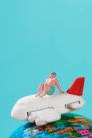 a miniature man, wearing swimsuit, sits on top of an airplane and a terrestrial globe, against a blue background simulating the sky with some blank space on topの写真素材