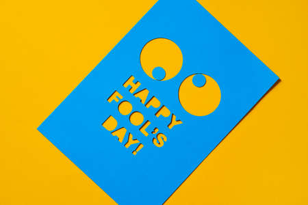 the text happy fools day and a pair of funny eyes cut out in a blue paperboard, against an orange backgroundの写真素材