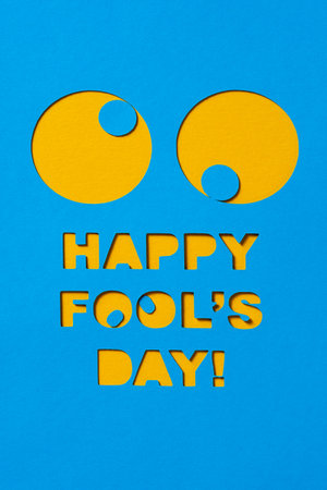a pair of funny eyes and the text happy fools day cut out in a blue paperboard, against an orange backgroundの写真素材