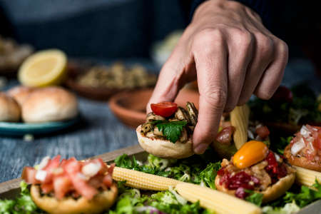 closeup of a young caucaisan man picking an appetizer from a wooden tray with some different vegan appetizers, with different toppings, placed on a gray tableの写真素材