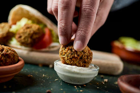 a young man dips a falafel in a bowl of yogurt sauce sitting at a rustic green wooden tableの写真素材