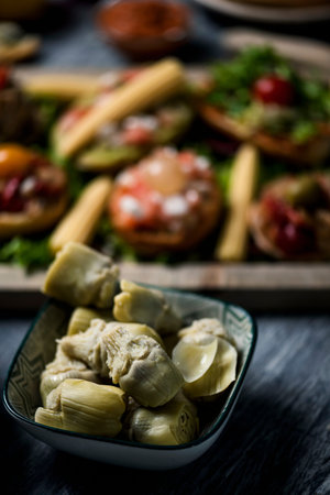 a rectangular ceramic bowl with some cooked artichoke hearts on a gray rustic wooden table next to a wooden tray with some different vegan appetizers, made with toasted bread and different toppingsの写真素材