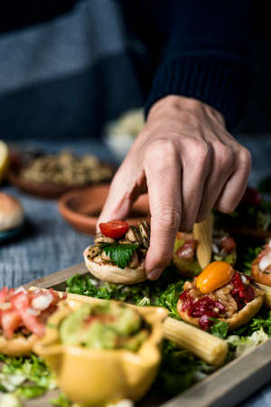a young caucaisan man takes an appetizer from a wooden tray with some different vegan appetizers, with different toppings, placed on a gray rustic tableの写真素材