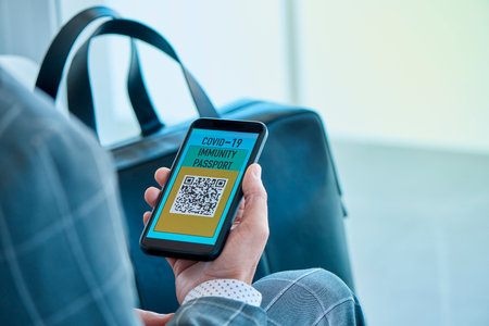 closeup of a young caucasian businessman, wearing an elegant gray suit, having a simulated electronic covid-19 immunity passport in his smartphone, sitting in the waiting hall of an airportの写真素材