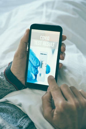 a young caucasian man wearing pajamas, lying in bed, checking the result of his covid test in a simulated app in his smartphone, that shows a negative result and reads covid test resultの写真素材
