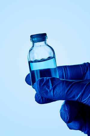 closeup of a healthcare or laboratory worker, wearing blue surgical gloves, having a vial with a blue liquid in his hand, against a pale blue backgroundの写真素材