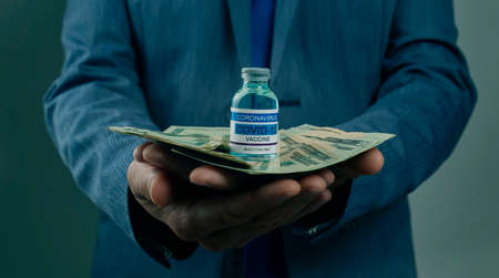 a young businessman has a simulated covid-19 vaccine bottle and a bunch of dollar notes in his handsの写真素材