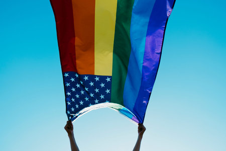 closeup of someone waving a rainbow US flag on the blue sky, with the sun in the background
