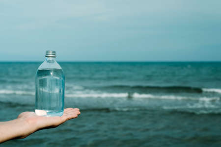 a young caucasian man has a glass reusable bottle full of water in his hand, in front of the oceanの写真素材