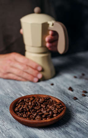 a young caucasian man is preparing some coffee in a beige moka pot, sitting at a gray rustic wooden table and some roasted coffee beans in the foregroundの写真素材