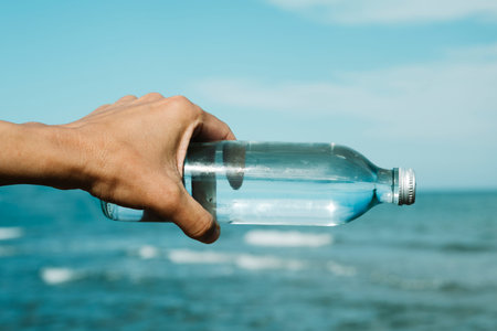 closeup of a young caucasian man holding a glass reusable bottle with water in his hand, in front of the ocean, aligned with the horizonの写真素材