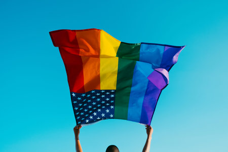 a young man, seen from behind, waves a rainbow US flag on the sky above his head