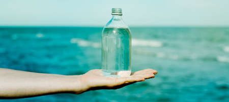 closeup of a young caucasian man holding a glass reusable bottle full of water in the palm of his hand, in front of the ocean, in a panoramic format to use as web banner or headerの写真素材