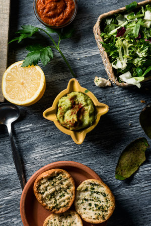 high angle view of a yellow ceramic bowl with some guacamole on a gray rustic wooden table, next to...