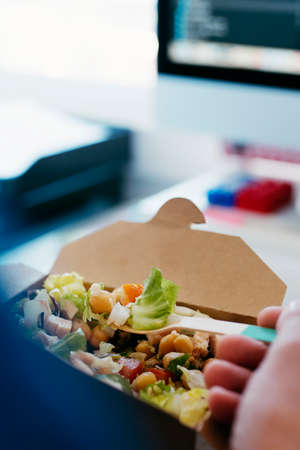 closeup of a young caucasian man eating a chickpea salad from a paper container, using a bamboo fork, sitting at his deskの写真素材