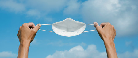 a young caucasian man holds a white FFP2 face mask in his hands against the sky, as he is taking a...