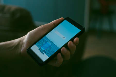 a young caucasian man, sitting at his living room, books his vacation online with his smartphone, with a simulated search engine in its screenの写真素材