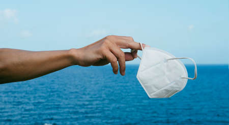 a young caucasian man holds a white FFP2 face mask in his hand in front of the ocean, as he is taking a breather of wearing it, in a panoramic format to use as web banner or headerの写真素材