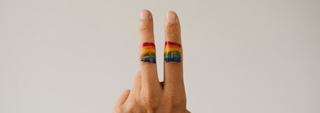 a rainbow flag in the fingers of a young person doing the V sign, against an off-white background with some blank space, in a panoramic format to use as web banner or headerの写真素材
