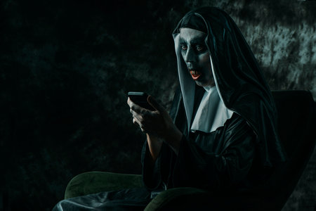 a scary evil nun, in a typical black and white habit, with bloody teeth, using her smartphone sitting in a chairの写真素材