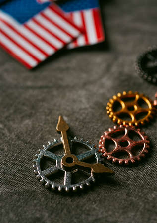 high angle view of some cogwheels, some clock hands and some small flags of the Uniteds States of America on a dark gray backgroundの写真素材