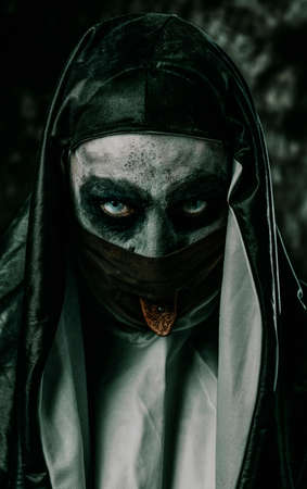 a scary evil nun, in a typical black and white habit, sticks out her pierced tongue through a hole in her black face maskの写真素材