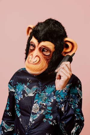 a young man wearing a monkey mask speaks in an old mobile phone, against a pink backgroundの写真素材