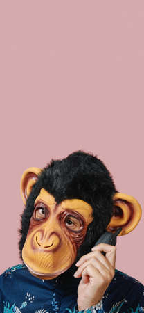 man wearing a monkey mask speaks in an old mobile phone, against a pink background with some blank space on top, in a vertical format to use for mobile stories or as smartphone wallpaperの写真素材