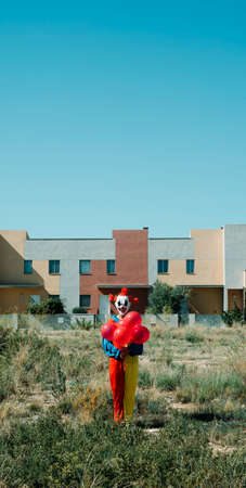 a creepy clown, wearing a colorful costume, holding a bunch of red balloons in his hand, standing in a vacant lot, in a vertical format to use for mobile stories or as smartphone wallpaperの写真素材