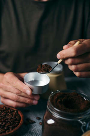 closeup of a young man filling the funnel of a moka pot with ground coffee from a glass jar, sitting at a gray rustic wooden tableの写真素材