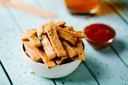 closeup of some fried strips of mock chicken in a bowl placed on a blue table next to a small bowl with ketchupの写真素材