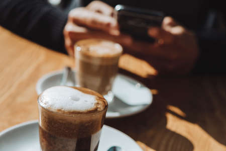 closeup of a young caucasian man using his smartphone, while is having a cappuccino, sitting at a table in an outdoors terrace of a sidewalk cafeの写真素材