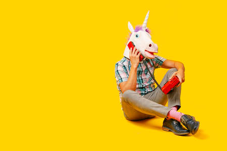 a young man wearing a unicorn mask talking on the phone, using a colorful red landline telephone, sitting on a yellow backgroundの写真素材