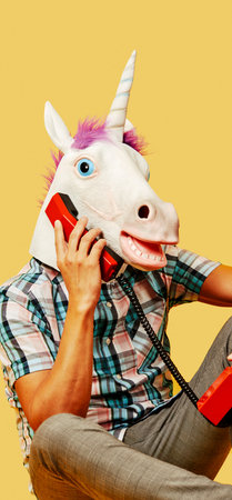 a young man wearing a unicorn mask talking on a colorful red landline telephone against a yellow...