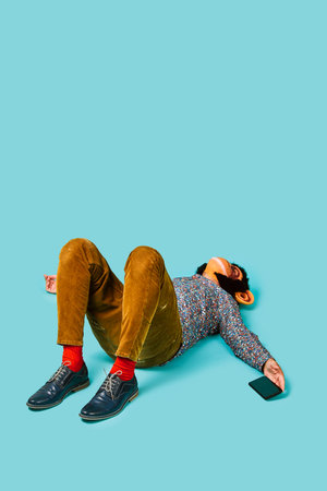 a young man, wearing a monkey mask, with his smartphone in his hand is lying down on a blue...