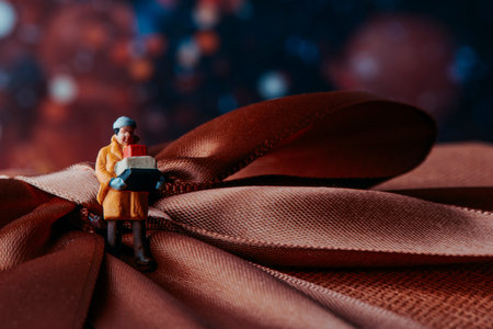 closeup of a miniature man, wearing a warming coat, carrying a pile of boxes standing on top of a gift box tied with a satin brown ribbon, in a bokeh effect backgroundの写真素材