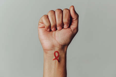 the raised fist of a man with a red awareness ribbon for the fight against AIDS painted in his wrist, against an off-white background with some blank space around himの写真素材